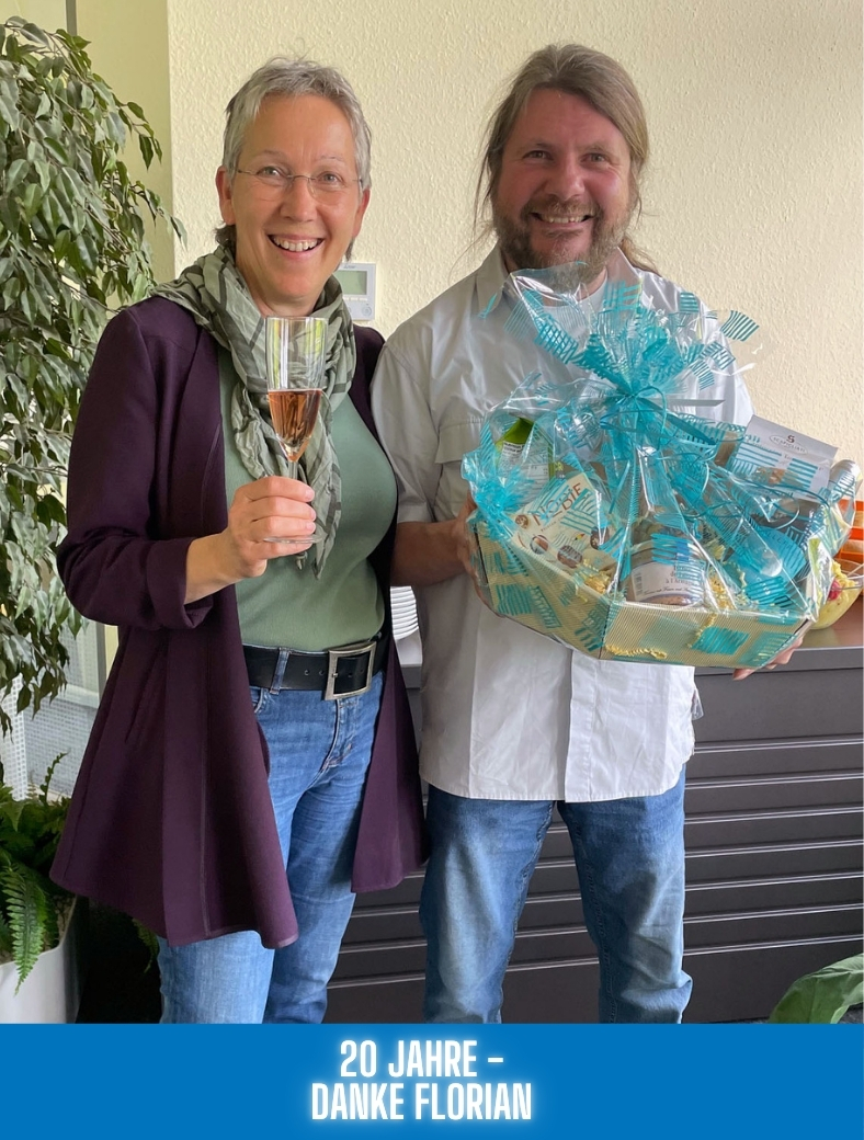 Geschäftsführerin Liane Schmauser mit Mitarbeiter Florian Arend lachend mit Geschenkkorb und Sekt zum anstoßen